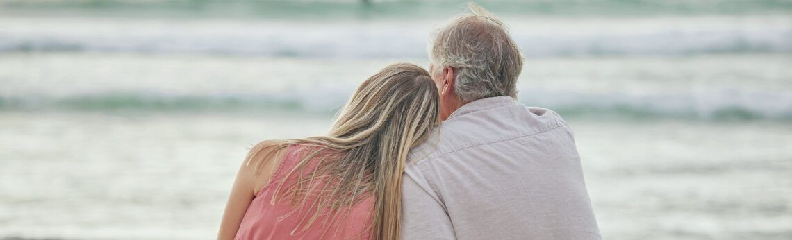 Couple embracing, watching the ocean. Blonde hair, pink dress, white shirt. Beach setting with waves.