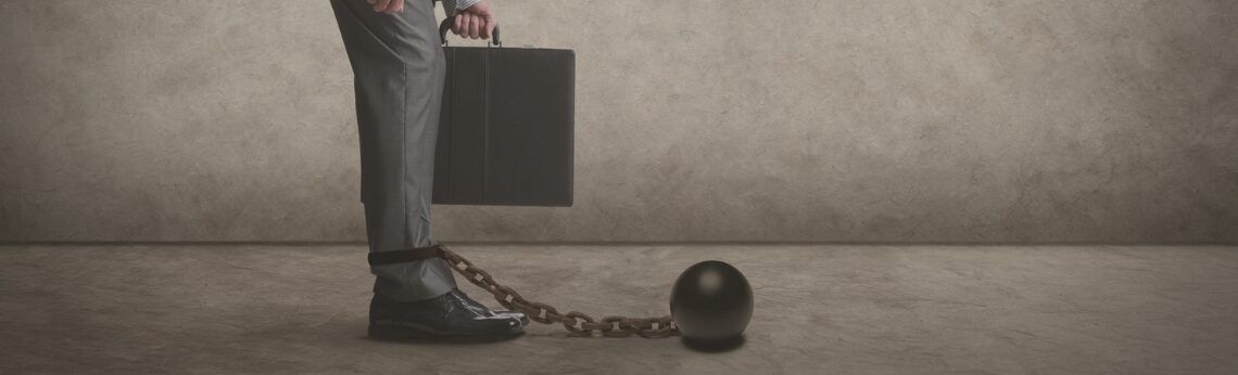 A person in a suit is chained to a ball, holding a briefcase.