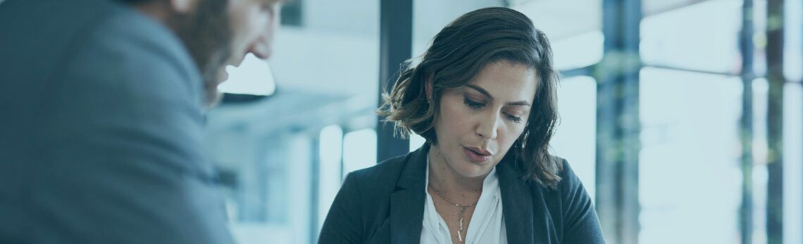 Woman in a blazer talking with a person, possibly in an office setting.
