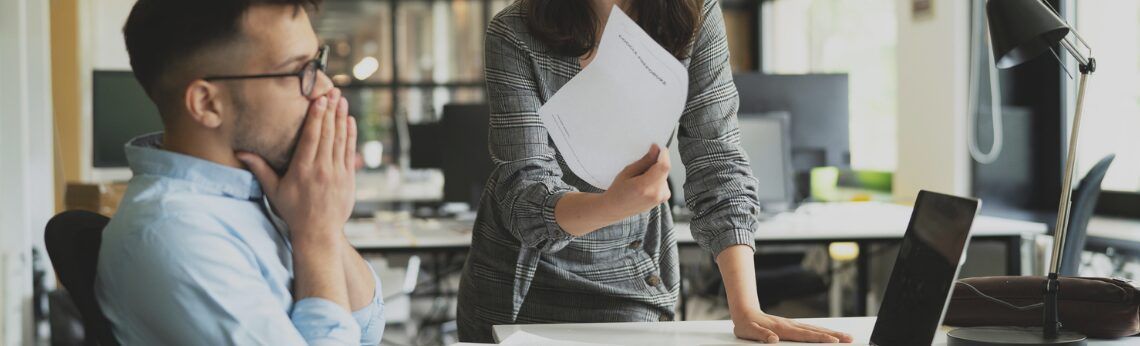 Man with hands covering face, looks stressed. Woman holds papers, leans on a desk in an office.