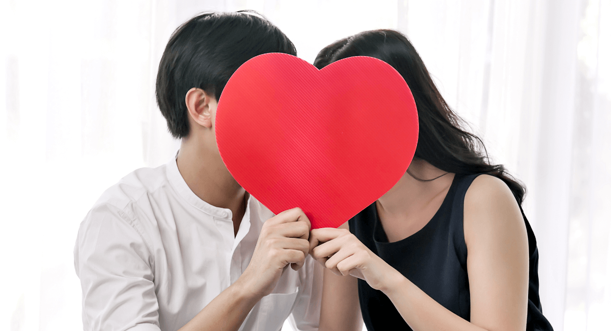 Couple holding a red heart in front of their faces, possibly kissing, against a bright background.
