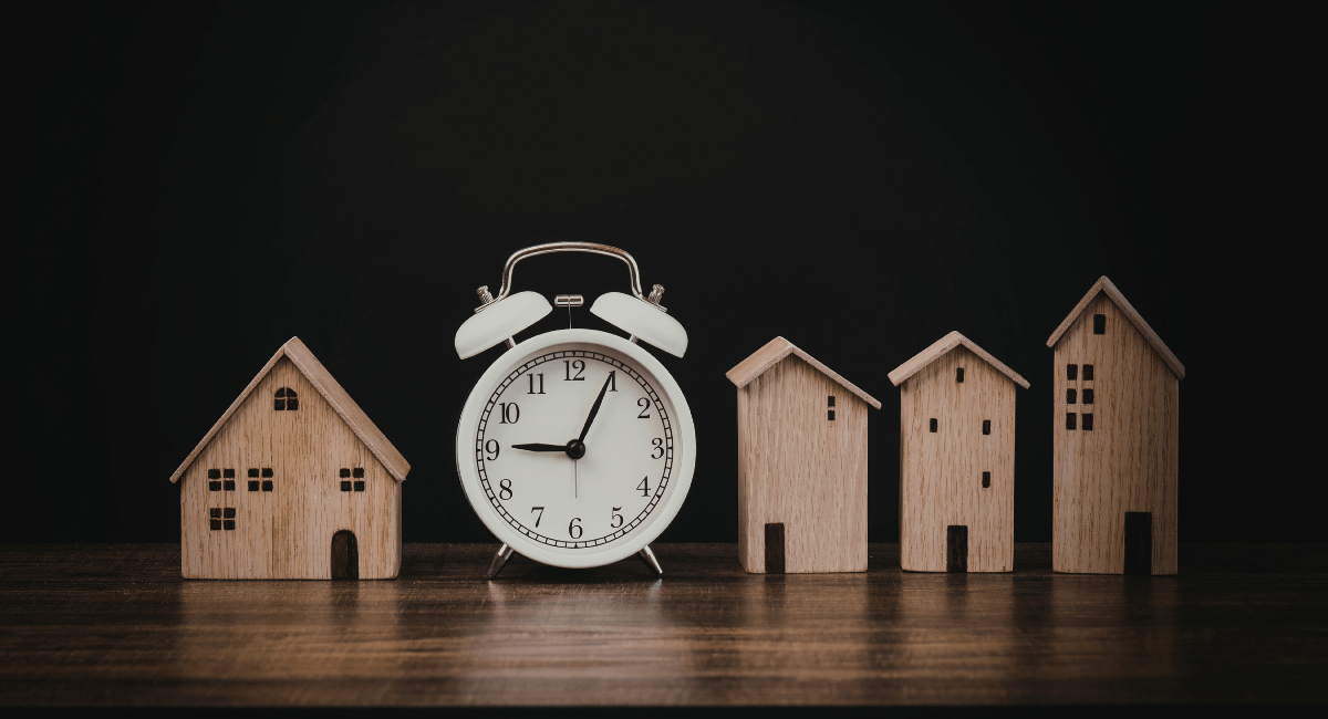 Clock and miniature houses on a table against a dark background, representing time and real estate.