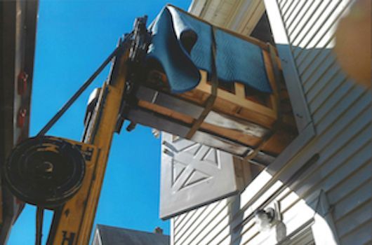 Yellow forklift lifting a wooden crate with blue cloth to a window of a building on a sunny day.