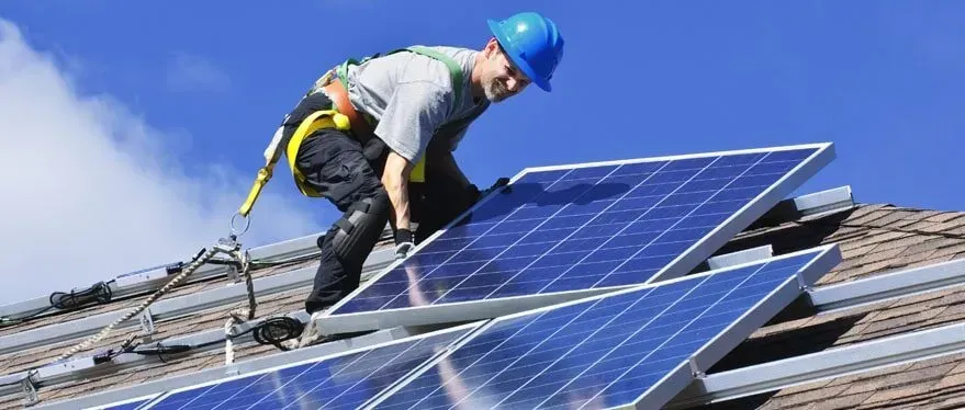 A worker in a blue hard hat installs solar panels on a roof. He is wearing a safety harness against a bright blue sky — AC Electrical & Air In Bundaberg Central, QLD