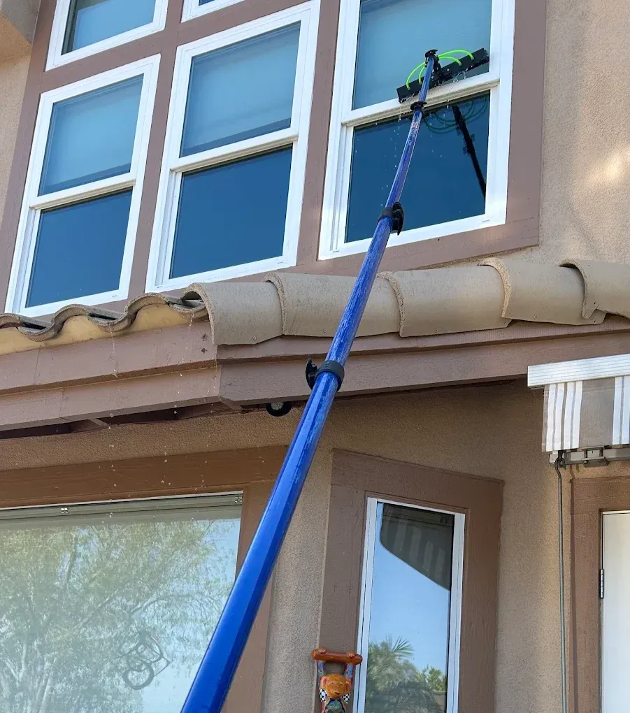 Person cleaning a second-story window with a blue extendable pole and squeegee. Brown trim, beige house, and blue sky.