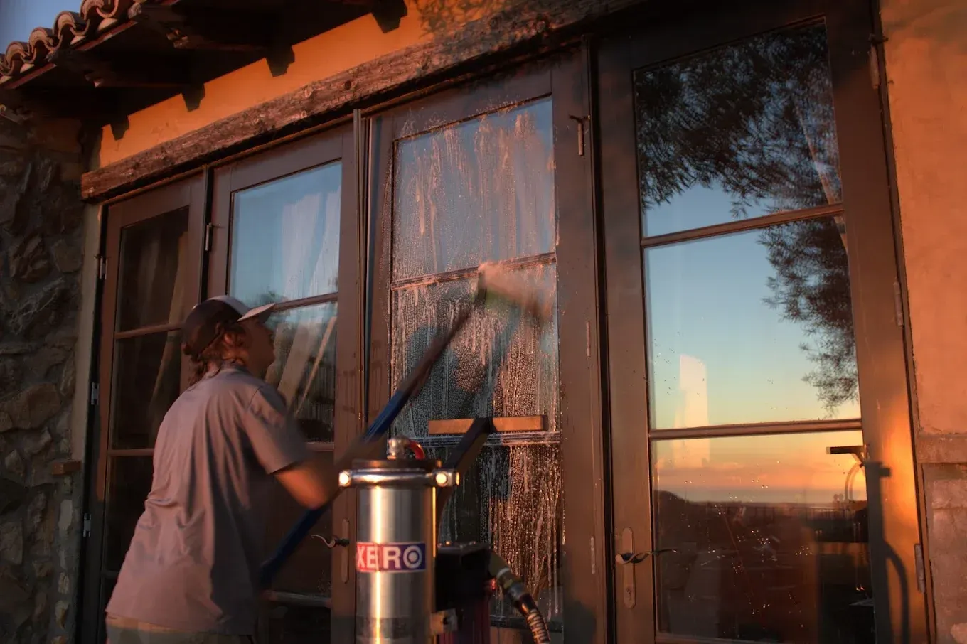 Person washing a window with a squeegee, next to a metal container. Sunset sky reflected in the glass.