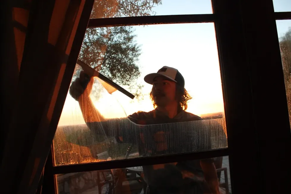 Man cleaning a window with a squeegee at sunset; warm light, outdoors.