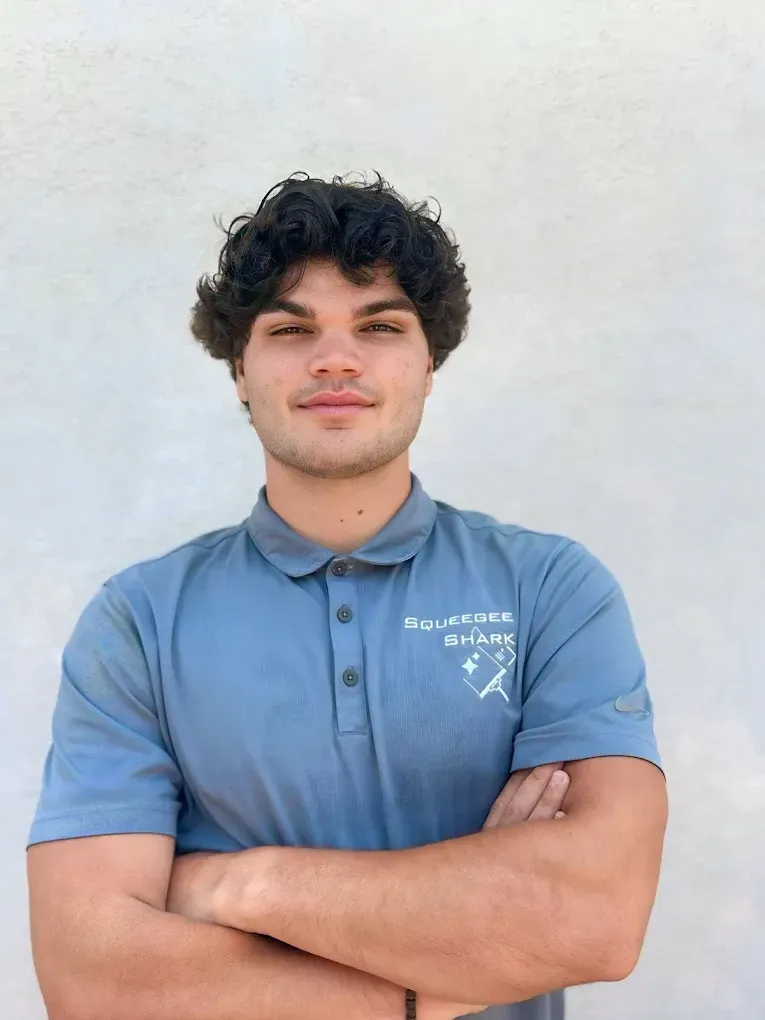 Young man with curly hair, arms crossed, wearing a blue polo shirt with logo, in front of a white wall.