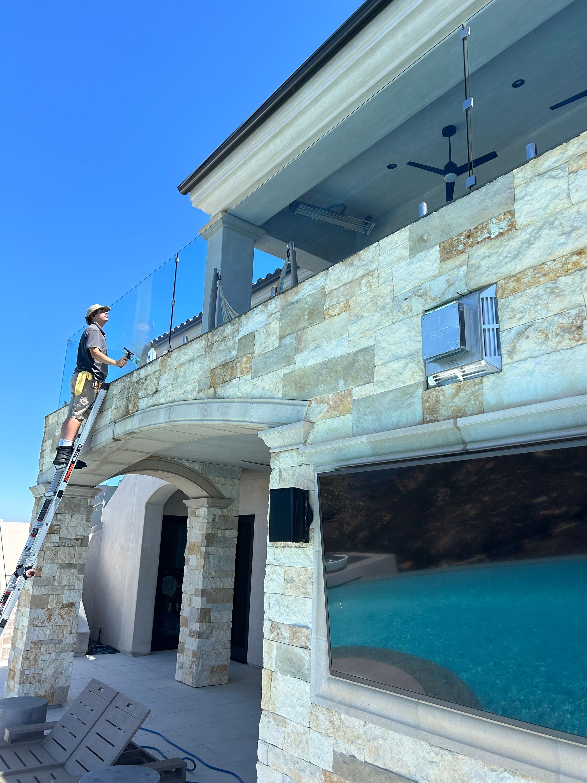 Person on ladder cleaning glass railing of a stone-walled building with an outdoor entertainment area.