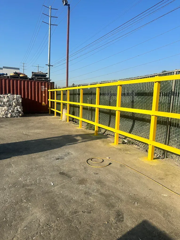 Yellow painted metal safety railing along a chain link fence. Industrial setting, bright sunlight.