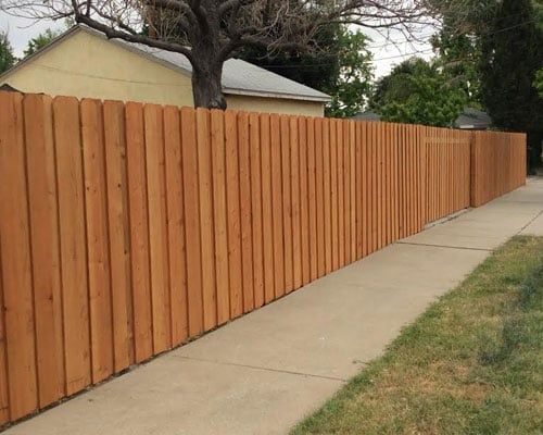 A wooden privacy fence along a sidewalk, with a house and tree in the background.
