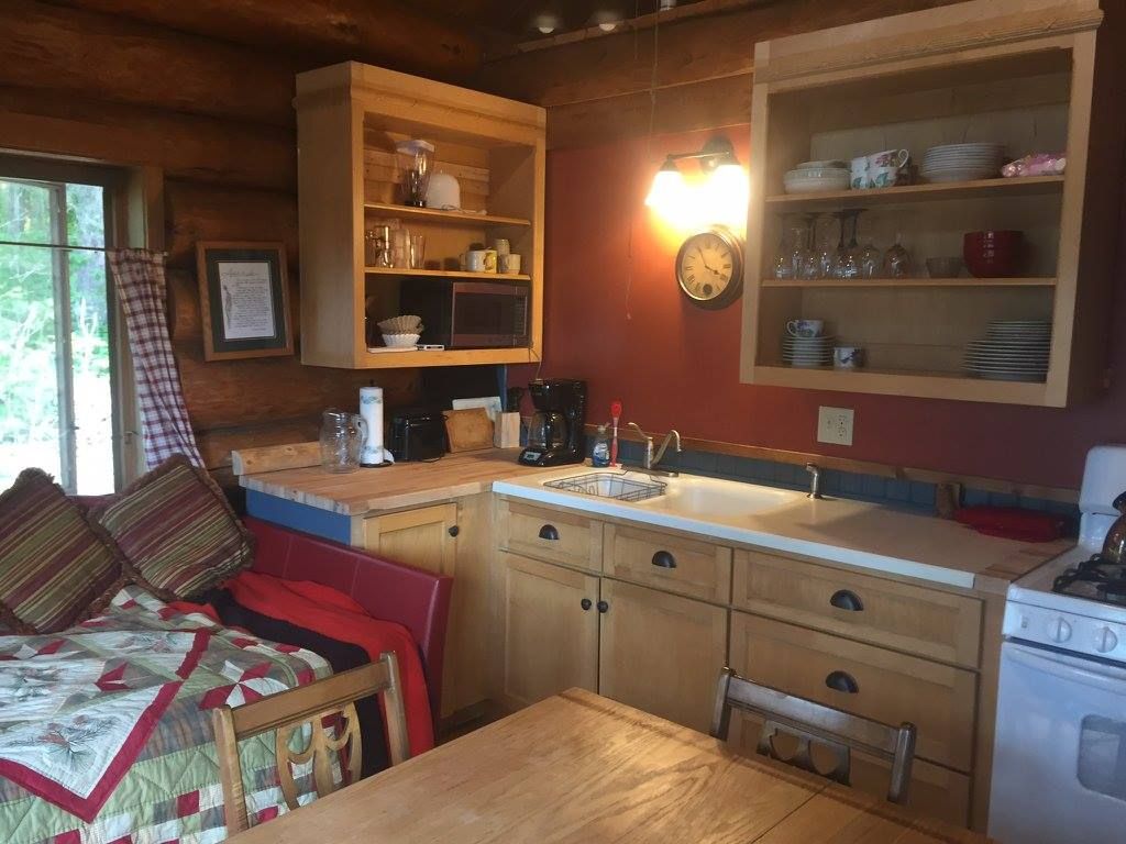 A kitchen in a log cabin with a table , sink , stove and shelves.