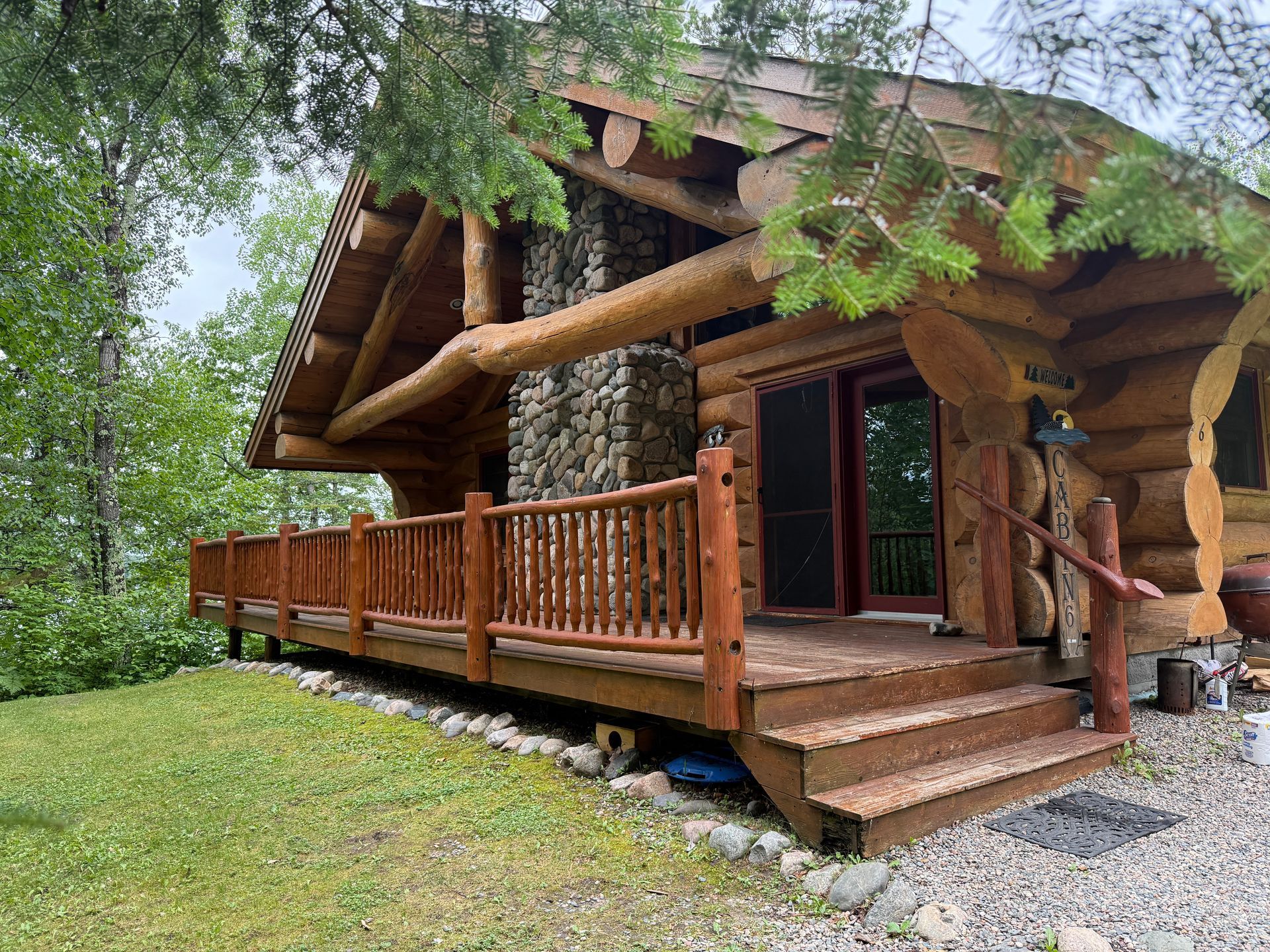 A log cabin with a wraparound porch, stone chimney, and wooden railings, nestled amongst trees. Brown logs and green foliage dominate.