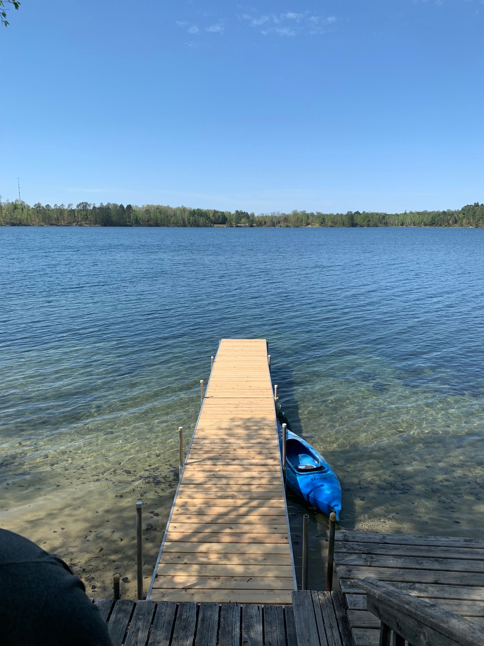 A wooden dock extends into a clear blue lake with a blue kayak tied to it. A clear, cloudless sky is overhead.
