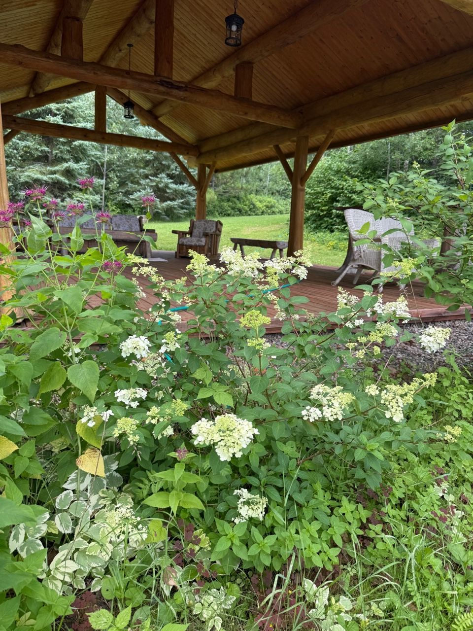 A wooden gazebo surrounded by lush green plants and flowers. Chairs and a table are inside the gazebo on a red brick floor.