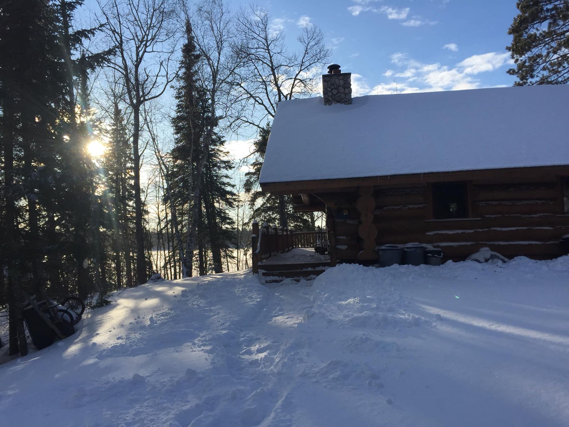 A log cabin is surrounded by snow and trees and the sun is shining through the trees