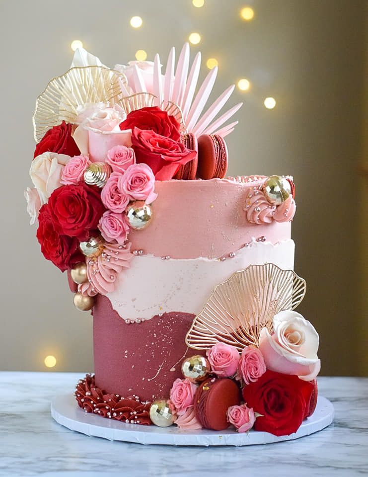 A pink cake with red roses and macarons on top is sitting on a table.