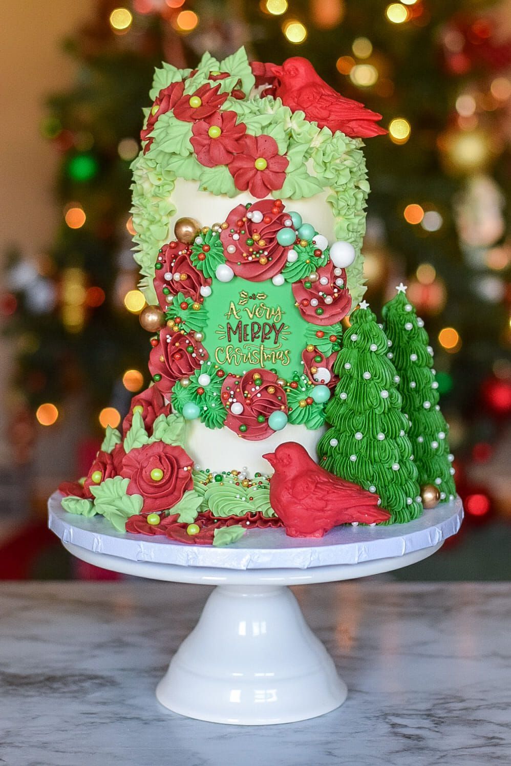 A christmas cake is sitting on a cake stand in front of a christmas tree.