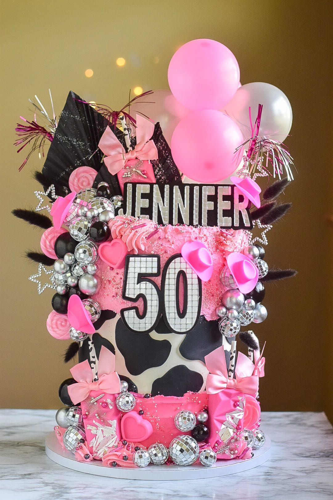 A pink and black cake with balloons and a cow print is on a table.