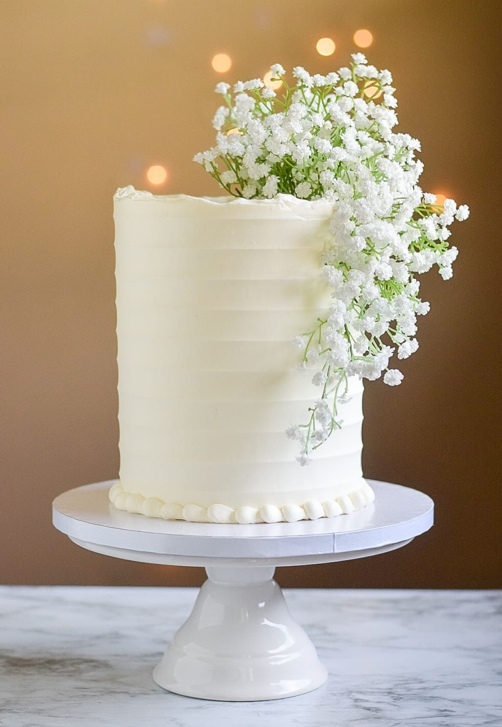 A white cake with baby 's breath on top is on a white cake stand.