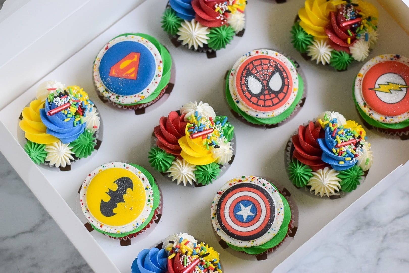 A box of cupcakes decorated with superhero logos and sprinkles.