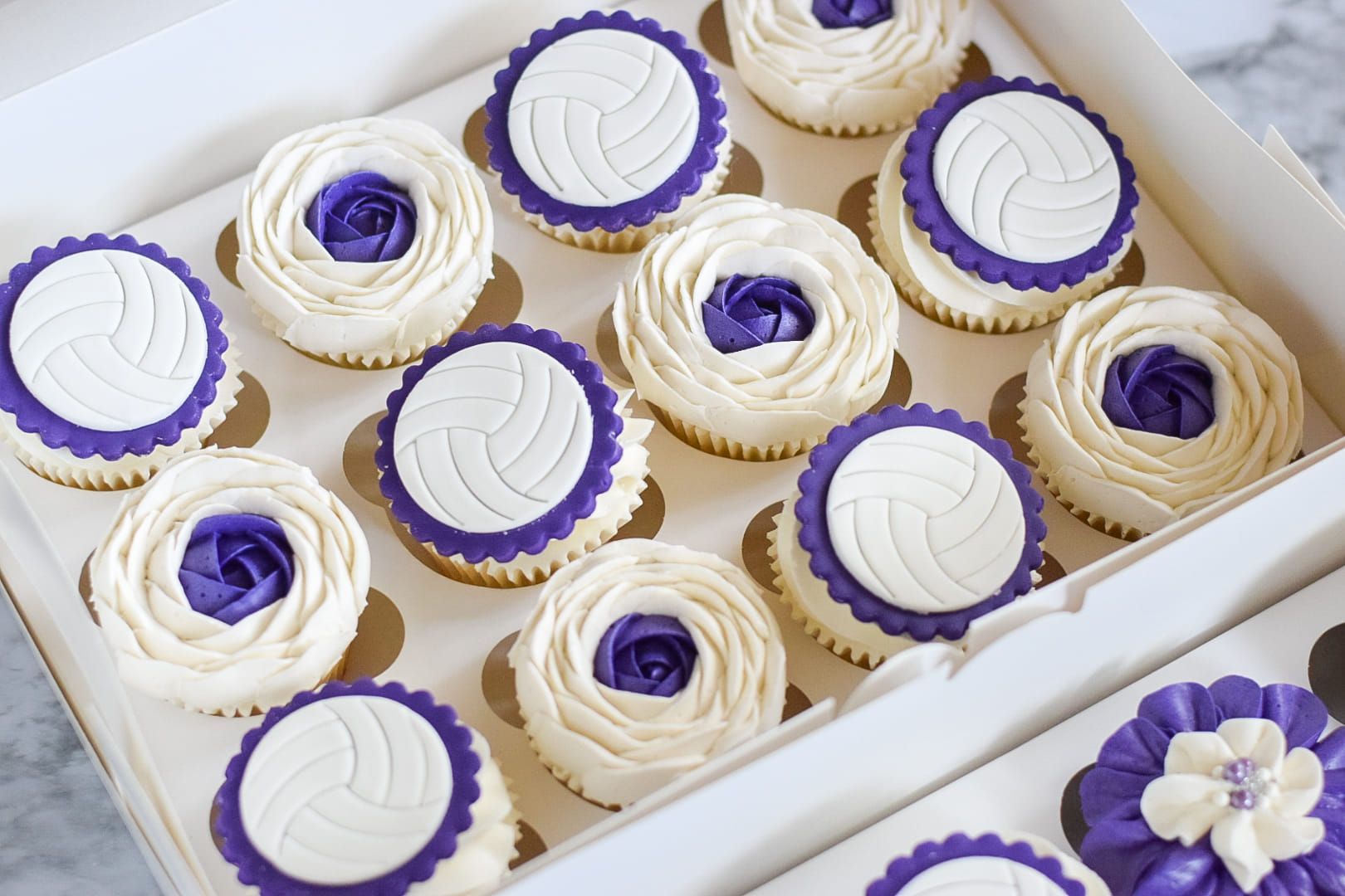 A box of cupcakes with purple frosting and volleyball designs on them.