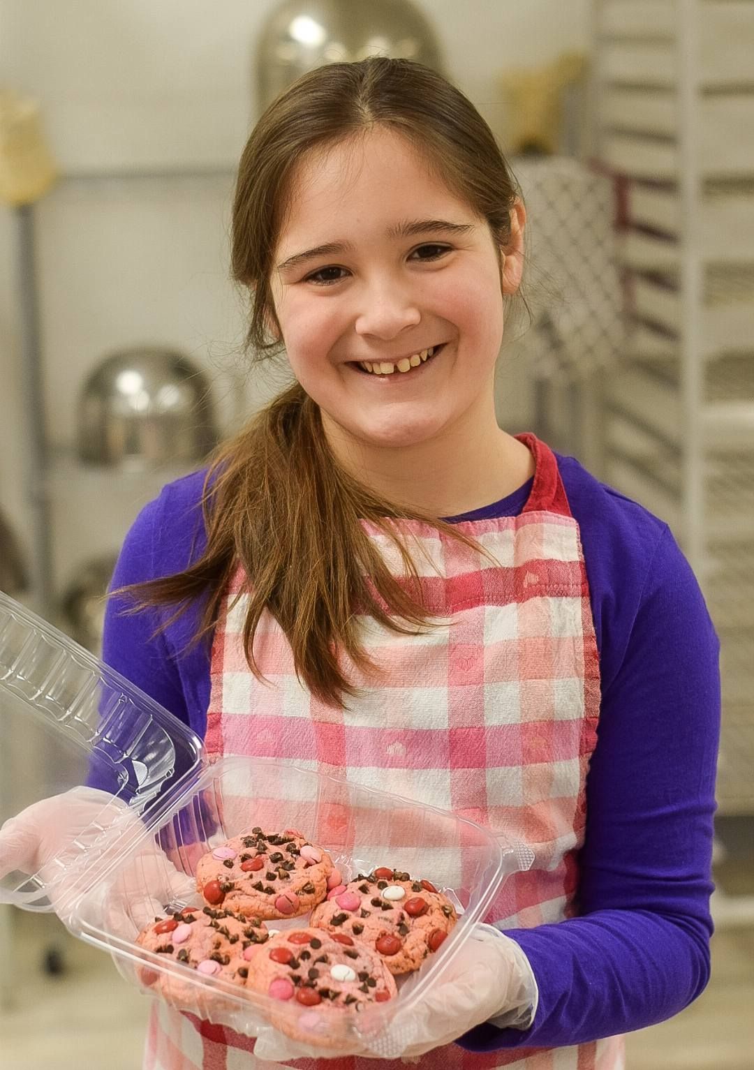 A young girl in an apron is holding a plastic container of cookies.