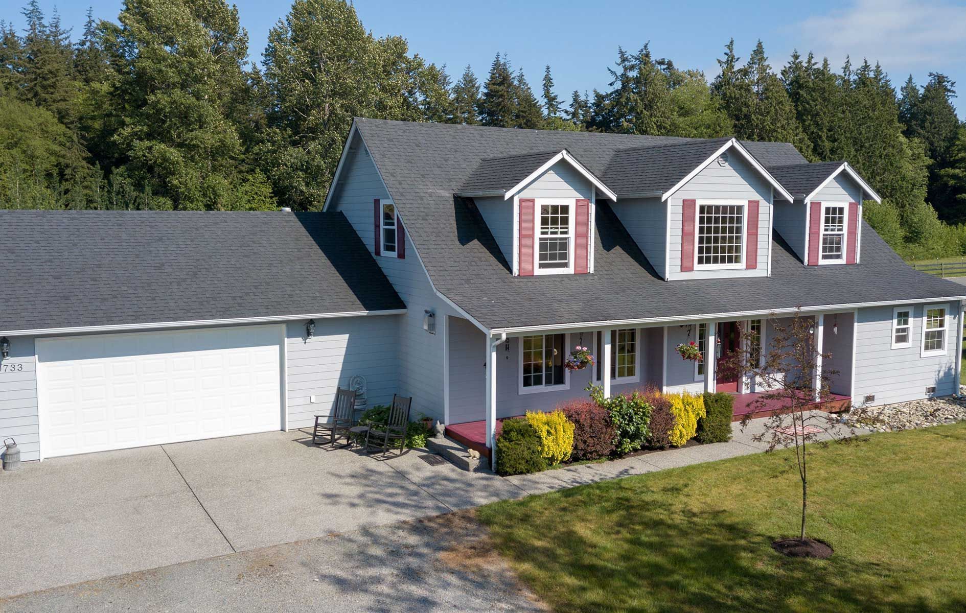 Suburban gray house with white garage, dark roof, and red shutters, viewed from the front yard.