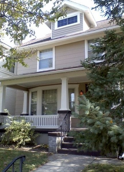 Two-story beige house with a front porch, white trim, and a large evergreen tree in the yard