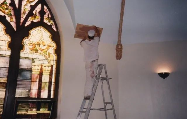 Worker on a ladder repairing a ceiling beside a stained-glass window in a church-like room