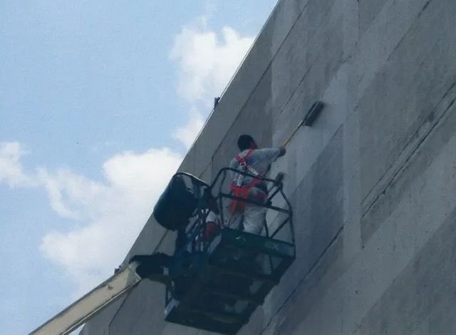 Worker on a boom lift painting a tall concrete wall under a blue sky.