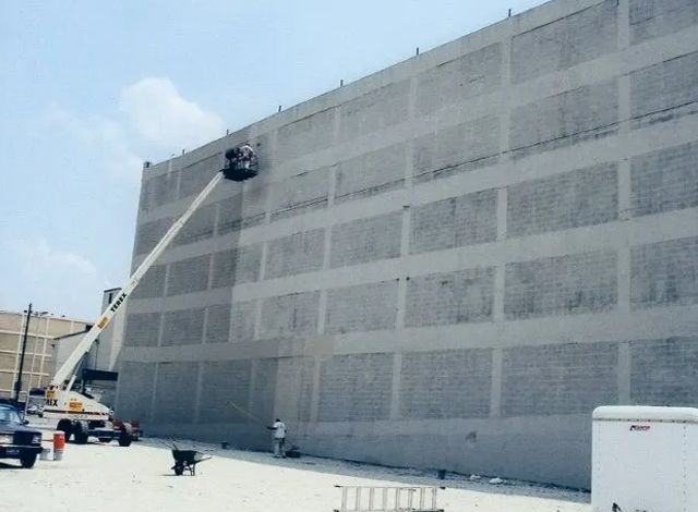 Worker in a lift painting a large gray industrial wall outdoors.