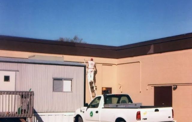 Worker standing on a white pickup truck beside a beige building under a blue sky