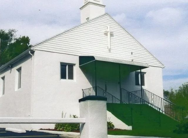 White church with a steep roof, green awning, and front staircase on a grassy hillside.