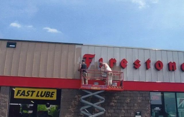 Workers on a lift installing red Firestone sign letters above an auto service store