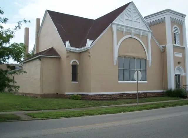 Tan church-like building with steep roof, arched windows, and a white-trimmed entrance by a street