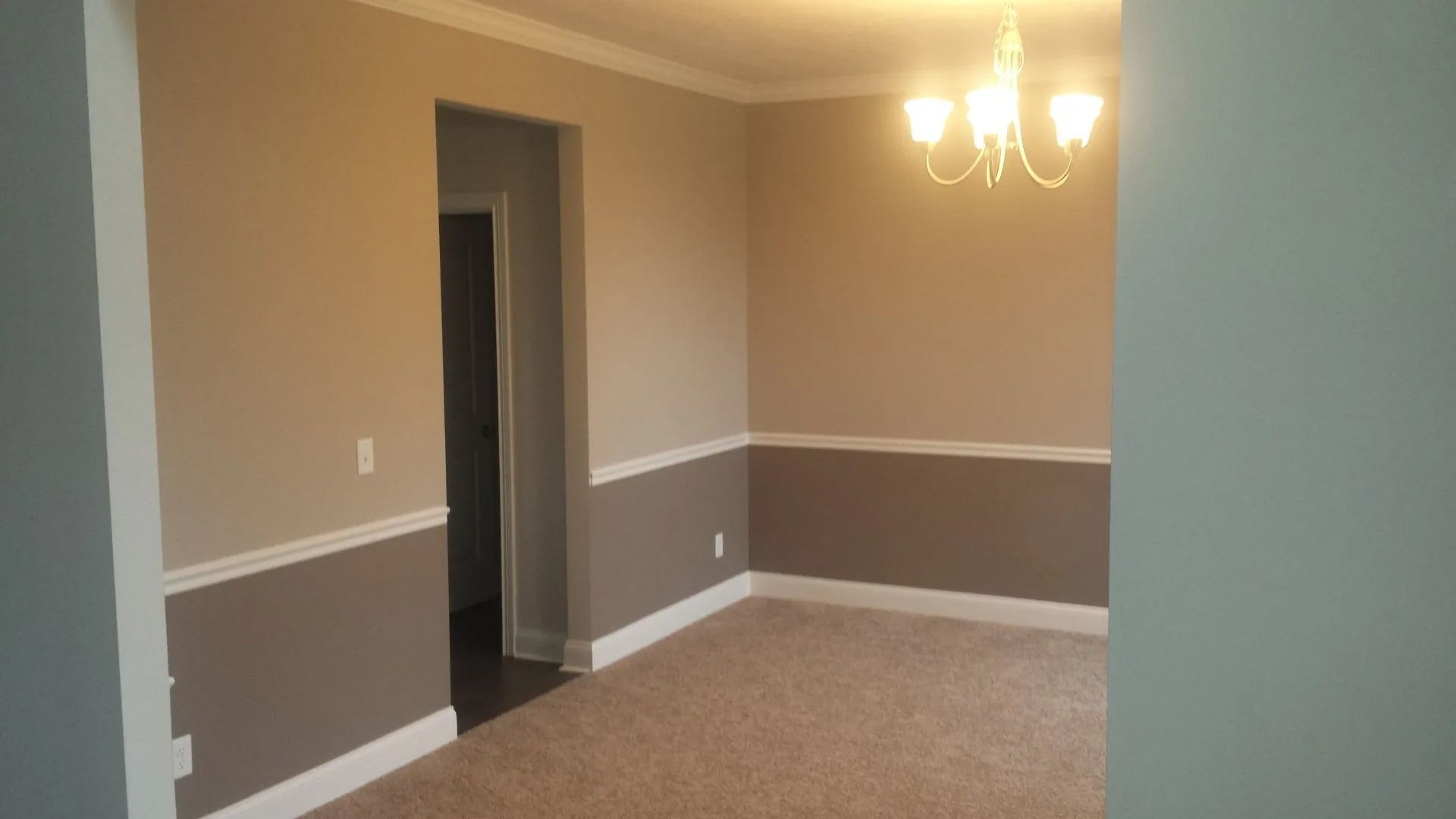 Empty beige dining room with brown chair rail, carpet, doorway, and chandelier light fixture
