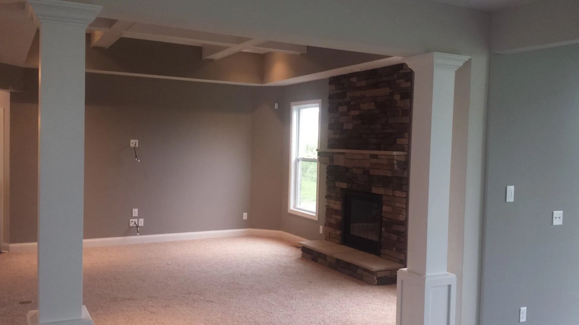 Empty living room with beige carpet, gray walls, and a brick fireplace by a window.