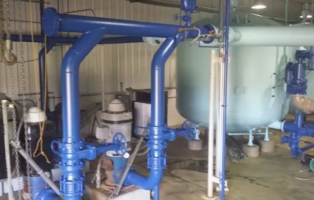Blue industrial pipes and valves in a mechanical room beside a large white tank.