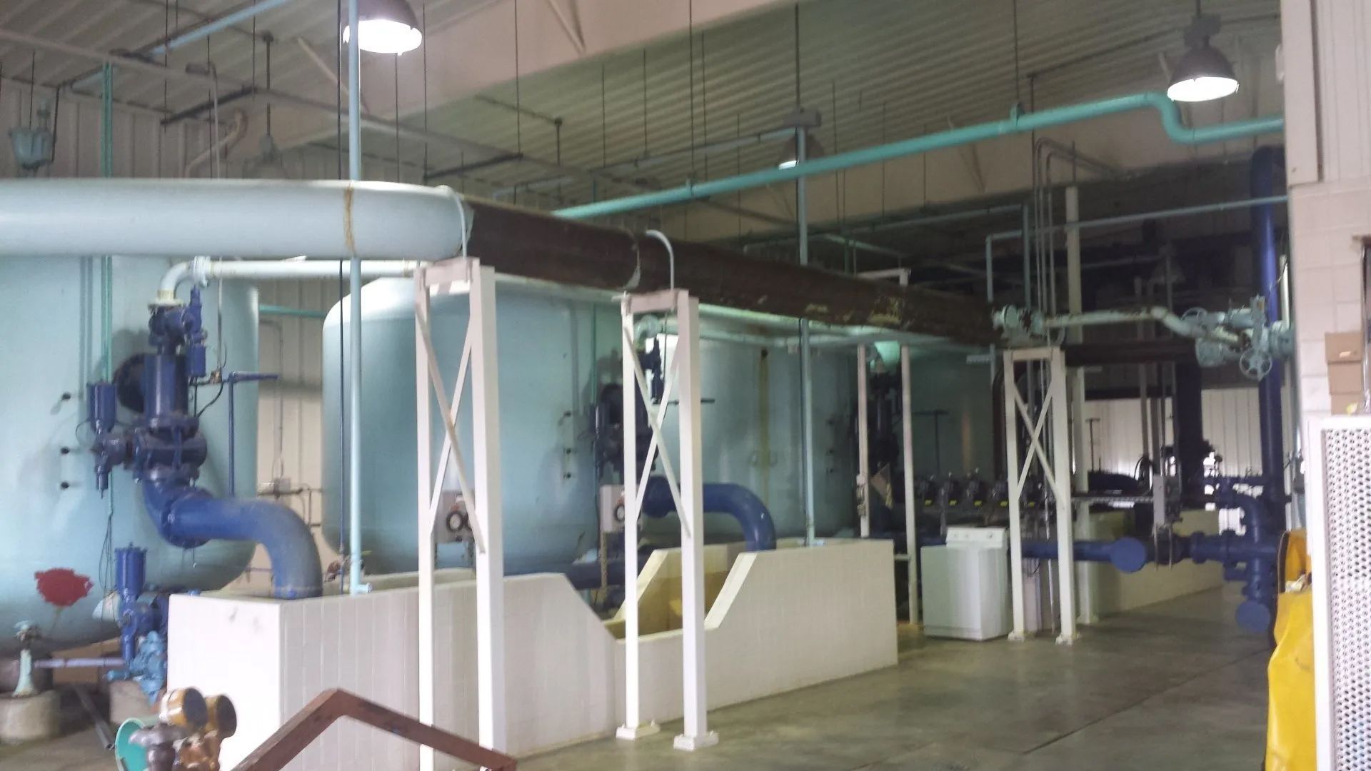 Industrial room with large blue and white pipes, tanks, and equipment under overhead lights