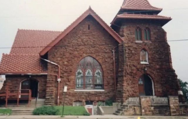 Brick church with a red-tiled roof and arched stained-glass window, viewed from the front