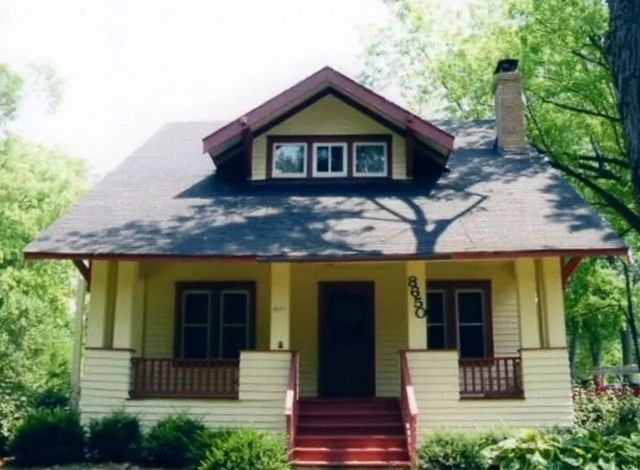 Yellow bungalow-style house with red steps, dark roof, and a front porch surrounded by trees