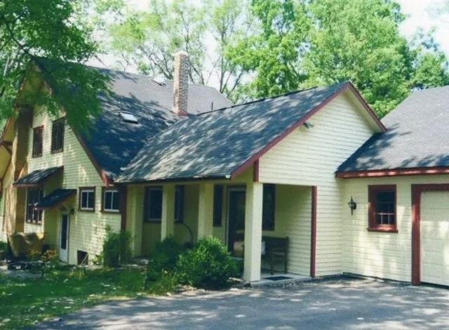 A cream-colored house with a dark roof and red trim, surrounded by trees and a driveway.