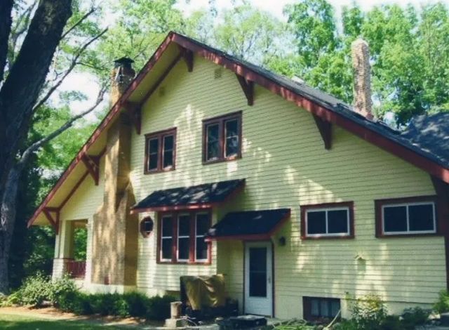 Green house with red trim and windows nestled among tall trees in a wooded setting