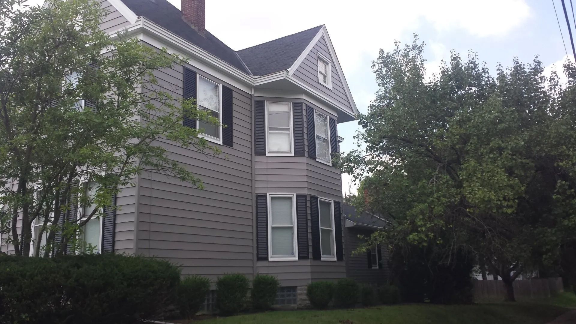 Gray two-story house with white trim and bay windows, partly framed by trees and greenery