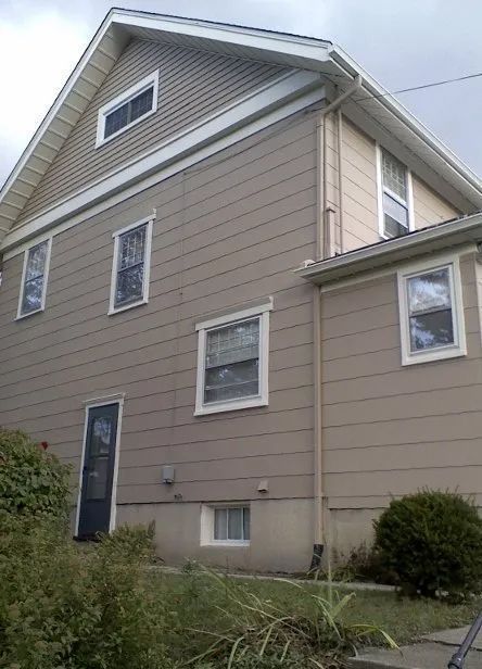 Side view of a beige two-story house with white trim and a small front bush.