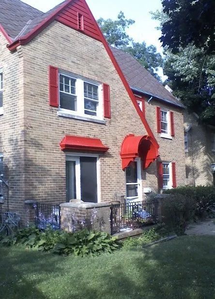 Brick house with red shutters and awnings, front lawn, and trees in the background