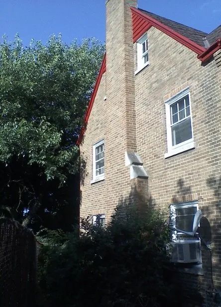 Brick house with red roof trim beside a leafy tree in bright sunlight