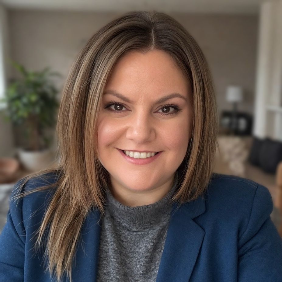 Woman with light brown hair smiles, wearing a blue blazer and gray turtleneck sweater indoors.