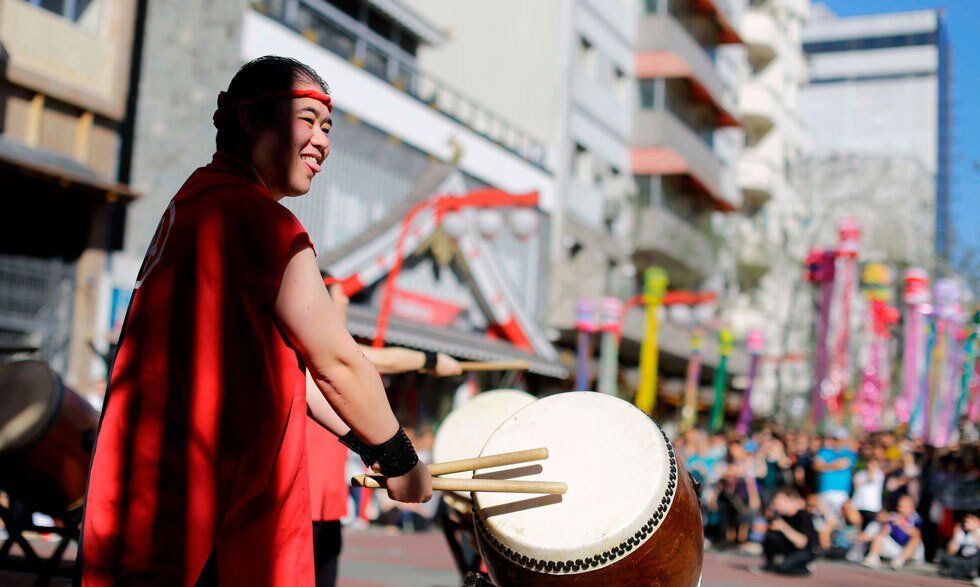 A smiling monk beats on a drum on a plaza while a crowd cheers